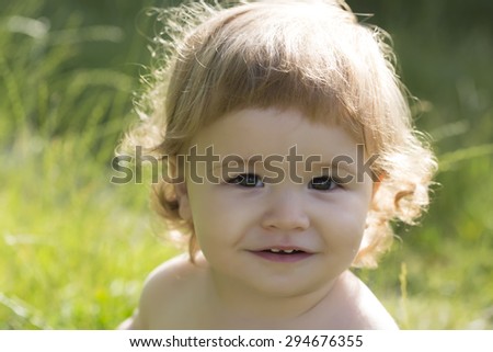 Portrait of little beautiful happy child boy with blonde curly hair smiling and sitting on fresh green grass outdoor on natural background, horizontal picture