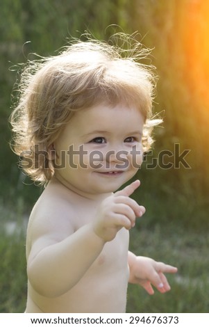 Funny playful cute smiling small baby boy with curly hair showing with finger standing outdoor sunny day on natural background, vertical picture