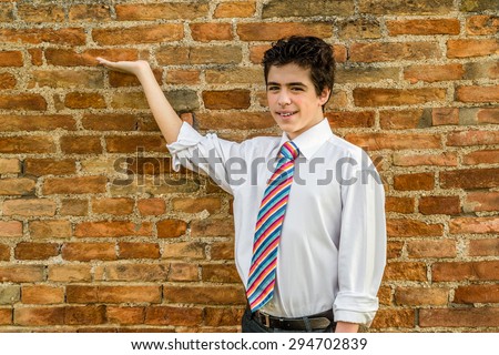 Handsome Caucasian boy wearing a white shirt and a regimental tie with red, fuchsia, orange, blue, indigo and white stripes is showing in front of a brick wall at sunset