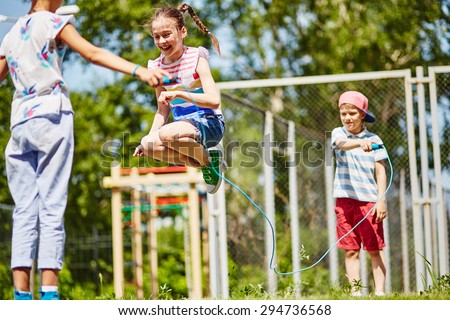 Happy girl jumping over skipping-rope held by her friends outdoors Royalty-Free Stock Photo #294736568