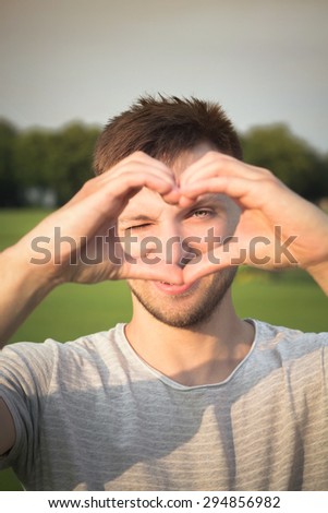 Young man making heart with his hands in beautiful sunset. Man looking at the camera through the heart.