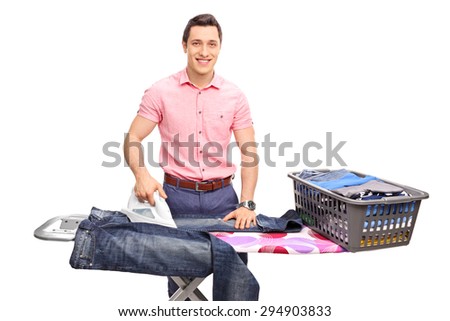 Cheerful young man ironing a pair of jeans on an ironing board and looking at the camera isolated on white background