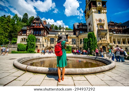 Female tourist in green dress, big hat and back pack photographing with digital tablet Peles castle in Romania. Back view