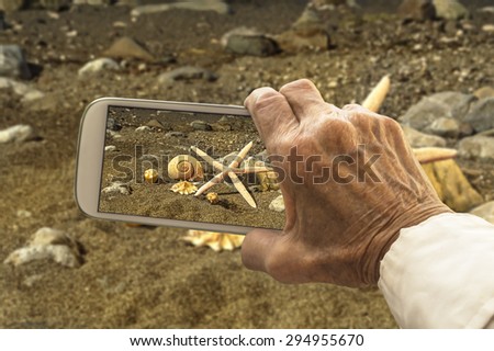 Old hand takes a picture of starfish and sea shells on smart phone. Selective focus.