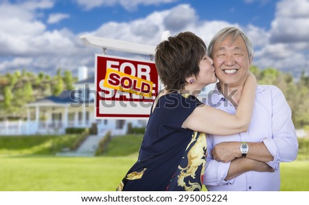 Attractive Affectionate Senior Chinese Couple In Front of Beautiful House and Sold For Sale Real Estate Sign.