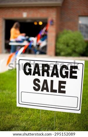 Garage sale sign on the front yard of a suburban home with a woman looking at items on a table.