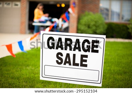 Garage sale sign on the front yard of a suburban house with a woman looking at items on a table.