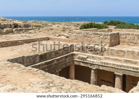Looking down into the grand chamber of an ancient carved stone burial tomb at an archaeological site in Paphos, Cyprus with the blue Mediterranean Sea in the background.