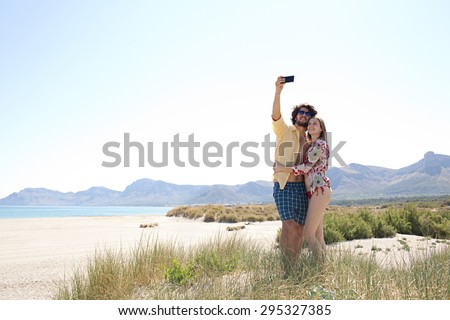 Young tourist couple on a white sand beach with dunes and mountains, enjoying a holiday, using a smartphone to take selfies photos of themselves, networking outdoors. Recreational travel lifestyle.