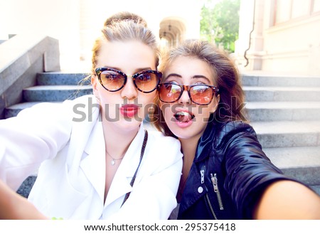 Close up lifestyle fashion portrait of two pretty fresh young brunette and blonde best friends girls,having fun,shows funny crazy faces,wearing sunglasses,bright jewelry,teenage outfit,urban,casual