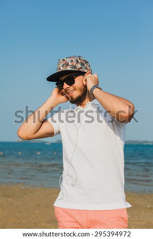  young guy in a baseball cap and sunglasses on the beach listening to music through headphones and smiling