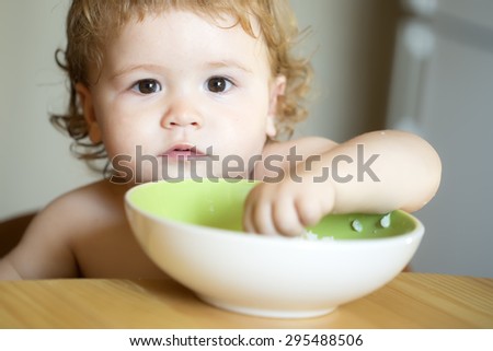 Portrait of interesting small pretty baby boy with blonde curly hair and round cheecks eating from green plate with hand closeup, horizontal picture