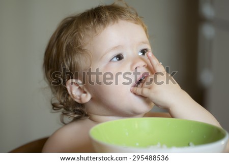 Portrait of curious small beautiful baby boy with blonde curly hair and round cheecks eating from green plate with hand closeup, horizontal picture