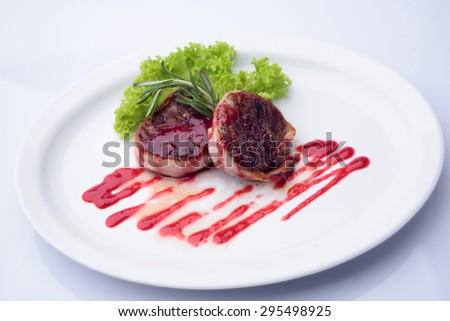 Pair of tasty grilled juicy pieces tenderloin wrapped with bacon drizzled with red sauce decorated with rosemary and lettuce on plate isolated on white, horizontal picture