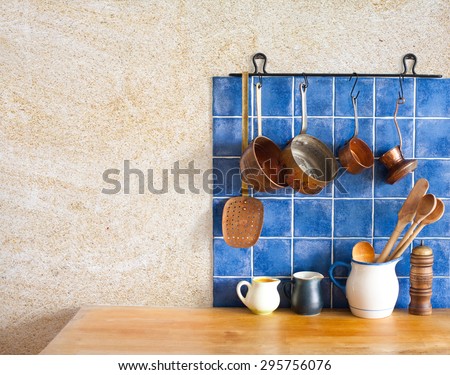 Kitchen interior. Hanging retro design copper kitchenware set. Pots, stewpots, coffee maker, spoon, skimmer. Different sizes, colors pitchers on the wooden table. Blue tiles, aged sand wall texture.