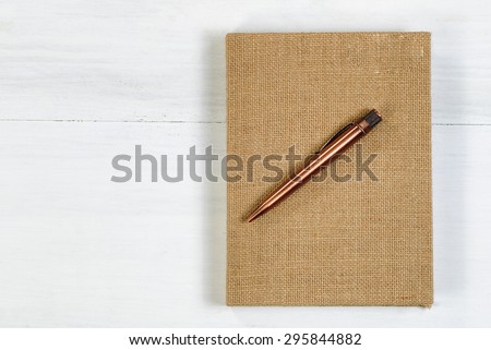 Vintage metal pen and burlap covered notepad on white wooden desktop.