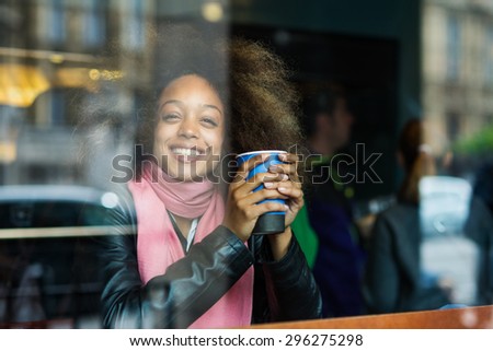 Smiling young woman portrait drinking coffee inside a cafe. Image shot through a window.