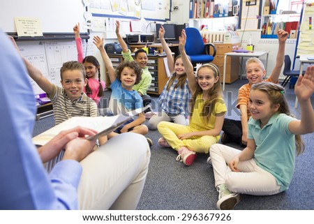 A group of children sit on the floor cross legged, listening to the teacher. They all have their hands raised in the air to answer a question.