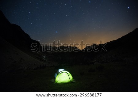 Lone, lit pop-up tent standing on a mountain pasture under the starry sky, night landscape with bright sun flare in the background. Outdoor lifestyle and sleeping under stars concept and background. 