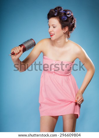 Young woman preparing to party having fun, amusing girl styling hair with curlers hairbrush retro style blue background