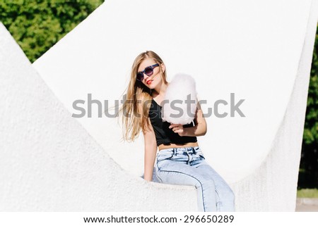 Portrait of beautiful blonde girl sitting outside with pink cotton candy. Wearing sunglasses. Red lips. White background, not isolated.