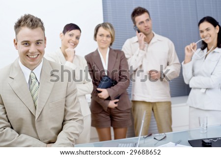 Young businessman sitting on office desk in front, looking at camera, smiling. Business team standing in the background.