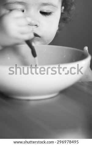Portrait of interesting little cute baby boy with curly hair and round cheecks eating from plate with spoon black and white closeup, vertical picture