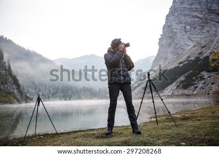 Photographer on assignment, holding a camera, taking photos of beautiful mountain landscape in the morning by a mountain lake with winter mist covered surface. 
