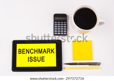 Business Term / Business Phrase on Tablet PC with a cup of coffee, Pens, Calculator, and yellow note pad on a White Background - Black Word(s) on a yellow background - Benchmark Issue