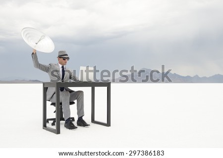 Businessman sitting working at his outdoor office desk trying to communicate holding up a satellite dish in stark white desert landscape Royalty-Free Stock Photo #297386183