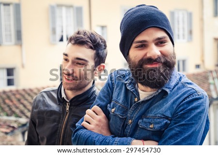 A pair of two young men take a break from work in the terrace of a building. One has a beard, hat and a shirt, the other a leather jacket