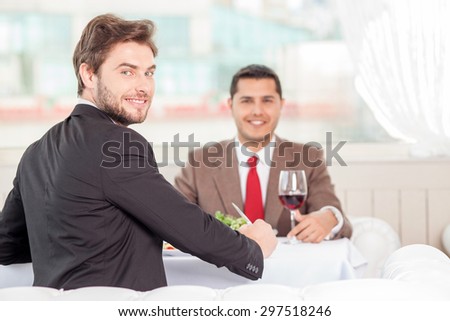 Cheerful male colleagues are sitting at the table in cafe. They are eating and laughing. The men discuss a contract and looking at the camera with joy. Copy space in right side