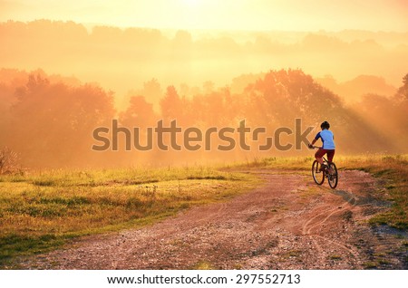 Young boy on the road sitting on his bike and watching amazing sunset with colorful rays during beautiful summer morning