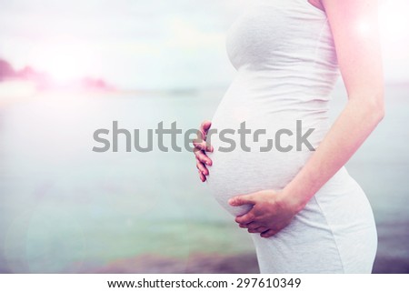 Close up profile view of the swollen belly of a pregnant woman cradling her unborn baby in her hands as she bonds, ocean sunset or sunrise background with copyspace Royalty-Free Stock Photo #297610349