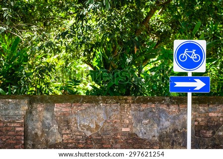 Bicycle sign post on the roadside with old brick wall and green trees