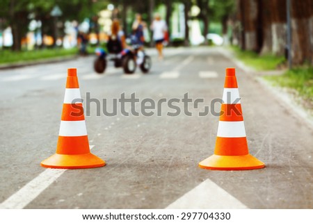 Closeup image of traffic  sign - orange cone with white stripes standing at at grey road at moving cars background.