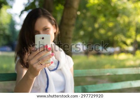 Young cheerful woman sitting on a Park bench in the shade of the trees and takes a picture on her smart phone. She holds a white phone in hand with red nail Polish. Holiday in the Park on a Sunny day.