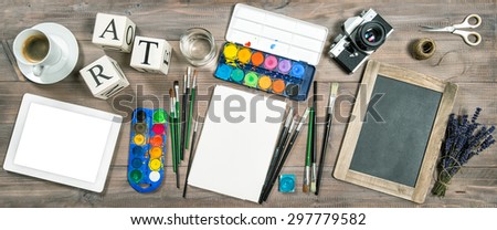 Artistic workplace mock up. Watercolor, brushes, digital tablet pc, chalkboard, vintage no name camera, office supplies, tolls and accessories. Retro style toned picture