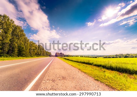 Rural road passing through fields and woods illuminated by the sun. View from the road, image in the orange-purple toning