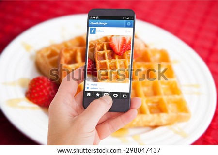 Female hand holding a smartphone against waffles and half cut strawberry together in a white plate