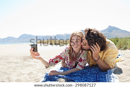 Joyful young tourist couple using smartphone to take selfies photo portraits, enjoying a summer holiday together, laying on sandy beach on vacation, kissing outdoors. Travel and technology lifestyle.