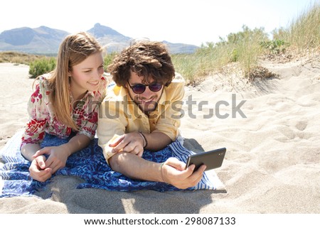 Portrait of a smiling young tourist couple using technology to take selfies photos, enjoying a summer holiday together, laying on a sandy beach on vacation, outdoors. Travel and technology lifestyle.