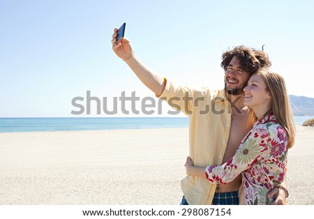 Portrait of young tourist couple on white sand beach, enjoying a summer holiday together, using smartphone to take selfies photos, networking outdoors. Recreational travel vacation lifestyle.