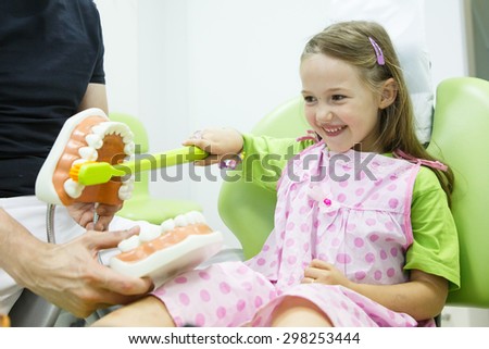 Smiling little girl in dentists chair, being educated about proper tooth-brushing by her paediatric dentist. Early prevention, raising awareness, oral hygiene demonstration concept.  Royalty-Free Stock Photo #298253444