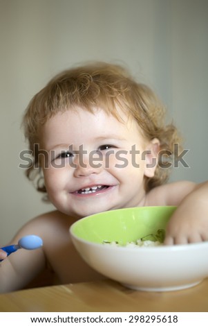 Portrait of laughing little baby boy with blonde curly hair and round cheecks eating from green plate closeup, vertical picture