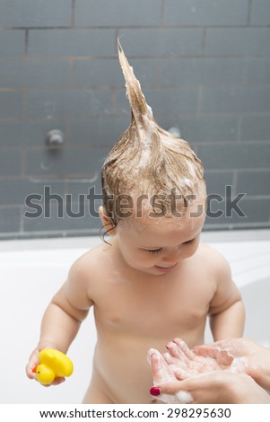 Small pretty male kid standing in bathroom with wet foam hair holding yellow duckling toy looking at hands of mother, vertical picture