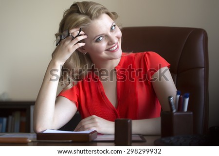 Pretty young elegant smiling business woman sitting in office on brown leather chair in red blouse holding glasses in hands looking forward indoor on white background, horizontal picture