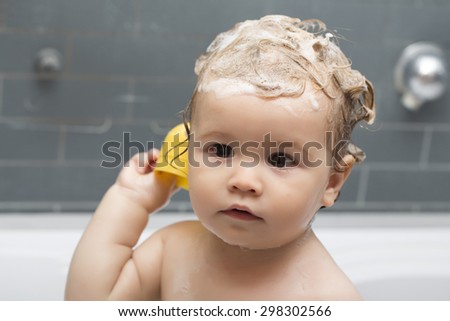Small pretty baby boy sitting in bathroom with wet foam hair holding yellow duckling toy looking away, horizontal picture