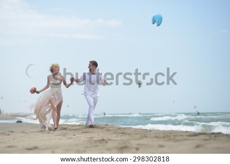 Pretty happy young wedding couple of boy and girl in white running along ocean beach coast on windy weather sunny day with paraplanes on blue sky background, horizontal picture