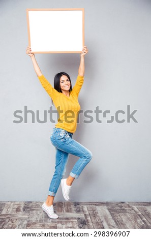 Full length portrait of a smiling woman posing with blank board on gray background